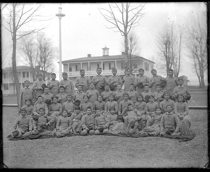 Picture shows Carlisle boys and girls in school uniform, dormitories in background from 1879