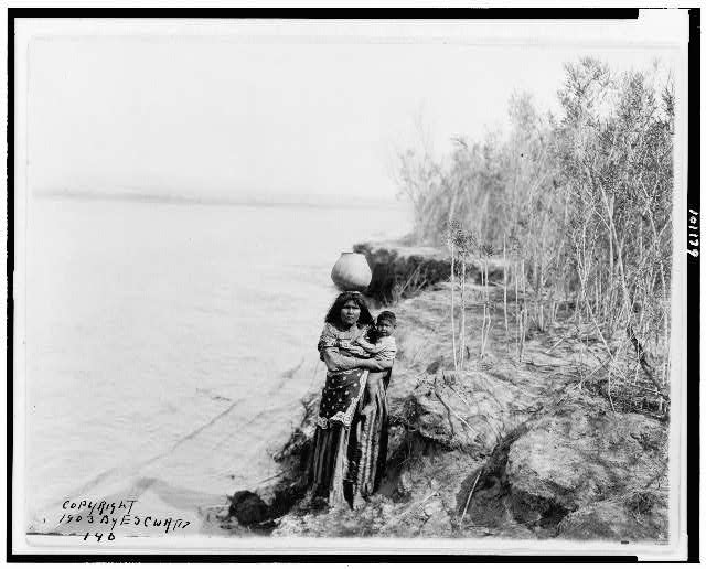 Picture shows a Mohave woman carrying water on her head and holding child in her lap from 1907