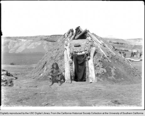 Navajo man sitting in front of his Hogan, ca. 1900. Hogan is in early style, constructed using the forked stick technique.